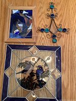 Photo showing three stained glass window hangers on wood surface: large square with birds, smaller rectangular with abstract glass, decorative metal with blue glass.
