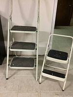 Front view of two metal step stools side by side showing three-step and two-step stools with black rubber steps and white paint splatters.
