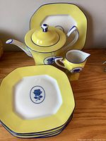Photo showing teapot with lid, creamer, and stacked plates on a wooden table with clear view of the blue rose design and yellow borders