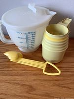 Photo of vintage Tupperware items on wood surface including a white measuring pitcher, four stackable yellow cups and yellow tongs.