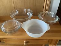 Photo showing four baking ware pieces on wood surface: clear glass bowls with floral and plain designs, clear glass casserole with lid, and white opaque two-handled ceramic/pyroceram dish.