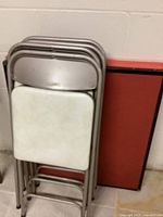Photo of 4 metal folding chairs stacked, showing padded white vinyl seat and back with gray metal frames, leaning against two folded vintage card tables with red tops and black edges.