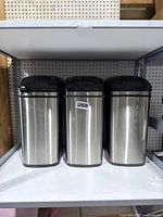 Three stainless steel battery operated automatic trash bins with black lids on a shelf.