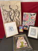 Photo showing all six art pieces arranged on a red fabric background. Includes large line sketch of three female figures, colorful graphic modern art, small pinup style mermaid, framed blue ink portrait, and other small framed artworks.