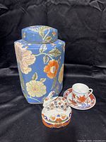 Full view of all four porcelain items: floral canister, rabbit trinket box, tea cup, and saucer against a black fabric background.
