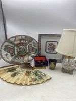 View of all items in the lot including platters, decorative folding fan, iron balls in box, small cup and desk lamp.