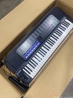Full view of the Casio Tone Bank CT-670 keyboard inside a cardboard box showing keys, speaker, microphone, and power cord.
