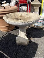 Full view of the cement bird bath showing the scalloped bowl and koi fish tail base outdoors on asphalt surrounded by pots and garden items.