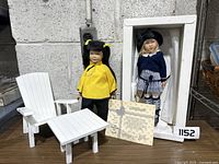 Two dolls displayed on a table alongside a white wooden chair and table set, with a certificate visible.