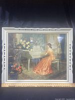Front view of framed Antoni Ditlef print showing woman in orange dress at white piano with floral decorations and elegant room background.