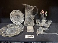 Full view of large glassware grouping including pitcher, decorative plates, small glasses, stirrers, and square container