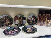 Photo of five M.J. Hummel collector plates arranged side by side along a shelf and the sealed collector book standing next to them, showing the detailed artwork on each plate.