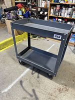 Full view of metal shop cart showing two shelves and caster wheels, placed in a workshop environment.