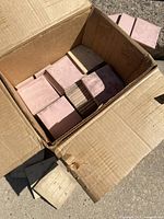 Opened cardboard box filled with stacked pink square bathroom tiles, approx 60 tiles visible. Box appears worn and dusty from storage.