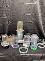 Image showing NutriBullet blender base surrounded by various transparent blending cups, blade attachments, and securing rings on black background.