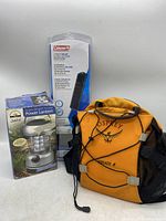 Front and side view of Osprey Helios 8 travel backpack in orange with Coleman solar battery maintainer and battery powered lantern in packaging behind it.