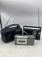 Three vintage portable radios grouped together. One is silver with cassette player and recorder buttons, another is a black radio with handle and antenna, and the third is a smaller black hand crank wind up solar radio flashlight with an antenna.