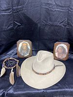 Full view of beige western style felt hat with leather strap band on black background with Native American style framed prints and dreamcatcher behind.