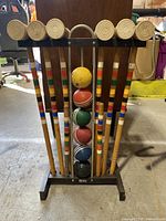 Front view of six wooden croquet mallets with colored stripes on handles, displayed on a stand with six croquet balls in various colors stacked in the center.