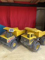 Front and side view of two vintage Tonka yellow dump trucks on a wooden surface against a red backdrop, showing their condition and size difference.
