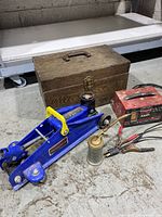 Wooden toolbox, blue hydraulic car jack, red battery charger, Eagle pump oiler on concrete floor.