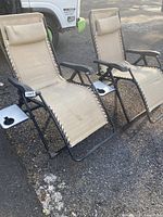 Pair of beige zero gravity chaise lounge lawn chairs with black metal frames and side trays with cup holders, showing overall weathered condition.