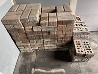 Stack of approximately 180 mixed solid and hollow red/brown bricks stacked in two neat piles with some hollow bricks distinguishable on the right side showing cylindrical holes and bits of concrete attached