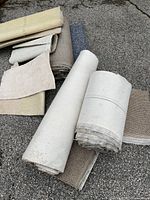 Multiple carpet remnants rolled or folded on ground, colours beige, white, light brown, blue. One white roll with small stain.