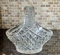 Clear diamond cut glass basket dish with handle shown from a side angle on dark reflective surface with brick wall background.