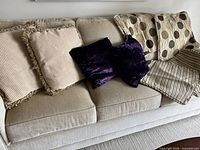 Four cushions arranged on a beige couch including two larger beige cushions with patterned covers and two smaller dark purple velvet cushions. Two sets of cushion covers in beige tones also visible.
