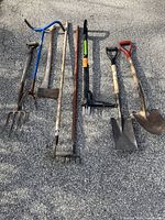 Full view of gardening tools laid on pavement including forks, hoes, shovels, axe, soil tiller, and Fiskars weed puller.
