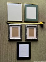 Top-down photo of five picture frames showing size comparison with measuring tape. Two gold-colored metal frames at top, three wooden frames with mats below.