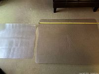 Both clear plastic chair mats placed on carpet with measuring tape showing size. Large mat on right approximately 60x46 inches, smaller mat on left approximately 49x35 inches.