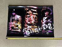 Poster laid flat partially rolled with a tape measure extended showing approximate 36 inch width, displaying three band members of Blink 182 playing instruments with purple and yellow stage lighting.
