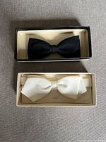 Photo showing two bow ties side by side in their boxes, one black and one beige.