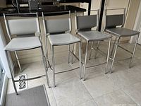 Four Bouclair bar stools in a row against kitchen cabinetry, showing grey faux leather seats and backs with metal frames.