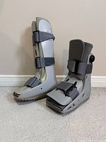 Pair of grey Aircast walking boots side by side on carpet. Each boot has hard plastic exterior support with three adjustable Velcro straps.