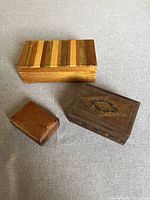 Three small boxes on a grey fabric surface: one large wooden box with striped slats lid, one dark wooden box with intricate lid design, and one small genuine leather box.