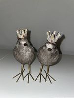 Front view of two gray bird figurines wearing gold crowns with red accents, standing on gold legs with three-pronged feet.