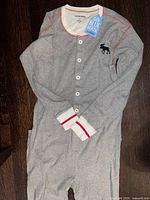 Front side photo of grey long-sleeve onesie with button closure, red-striped white cuffs, and moose logo on left chest.