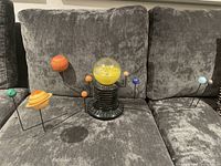 Solar system model displayed on grey couch showing colorful planets on wire arms orbiting central yellow illuminated sun dome on black base.