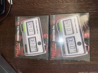 Two new Sealed Saitek Competition Pro game clocks in original black boxes placed side by side on wooden surface showing front of packaging with clock image and branding.