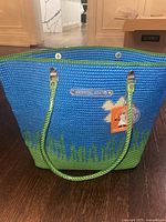 Front view of the large woven blue and green plastic beach bag with floral design and button closure, showing the Skipping Girl metal logo plate and green rope handles attached with metal hardware.
