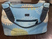 Front view of large blue floral fabric handbag showing large white, yellow, and blue daisy patterns, front zippered pocket, and black adjustable shoulder strap.