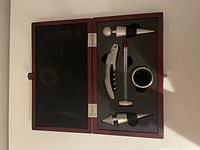 Open wooden box showing metal wine accessories placed in black foam interior: corkscrew, thermometer, two wine stoppers, and drip collar.
