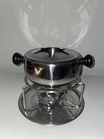 Stainless steel fondue set including pot with glass lid, mounted on metal stand with black handles visible.