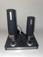 Front view of two black cylindrical wine warmers with silver bases on a black rectangular warming station with control dials and power adapter in the background.