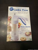 Front view of Wilton Cookie Press in box showing product image and key features including large 11 oz barrel, ergonomic handle, 12 disks, and 2 recipes