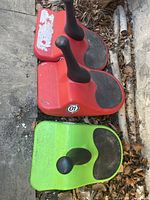 Three plastic Zipfy children’s toboggans, two red and one green, showing black handles, black foot grips, sitting outdoors on concrete or stone surface with leaves nearby.
