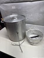 Front view of aluminum stock pot with lid, fry basket wrapped in plastic, and metal hook stand on table
